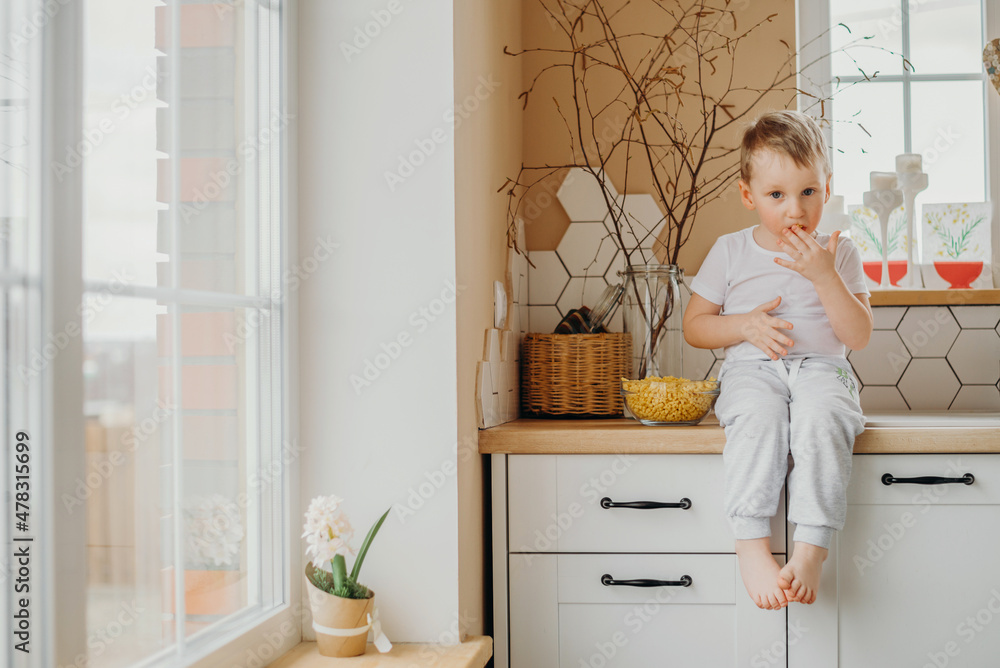 In the morning, a child, a boy, sits in the kitchen at home, on kitchen cabinets and eats fakes. It's spring outside, there's a crocus pot on the windowsill. In the morning, a child, a boy, sits in the kitchen at home, on kitchen cabinets and eats fakes. It's spring outside, there's a crocus pot on the windowsill.
