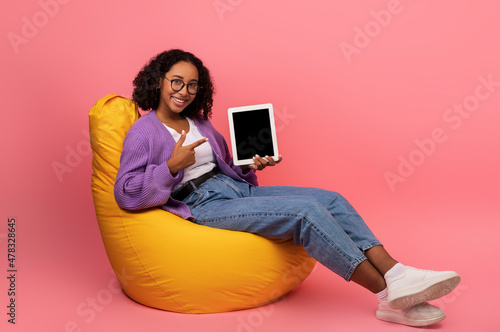 Young African American woman showing tablet pc with empty screen, sitting in bean bag chair on pink background, mockup