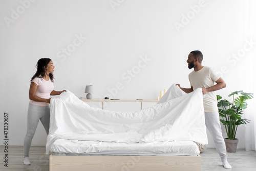 Fotografija Cheerful young black woman and guy make bed and hold blanket in bedroom interior