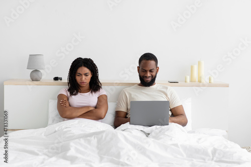 Offended sad young black woman angry at husband with computer, sit on bed in bedroom interior