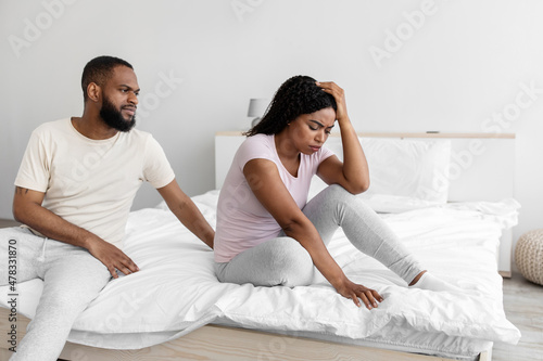 Serious young black man calm to sad desperate depressed wife, sit on bed in bedroom interior