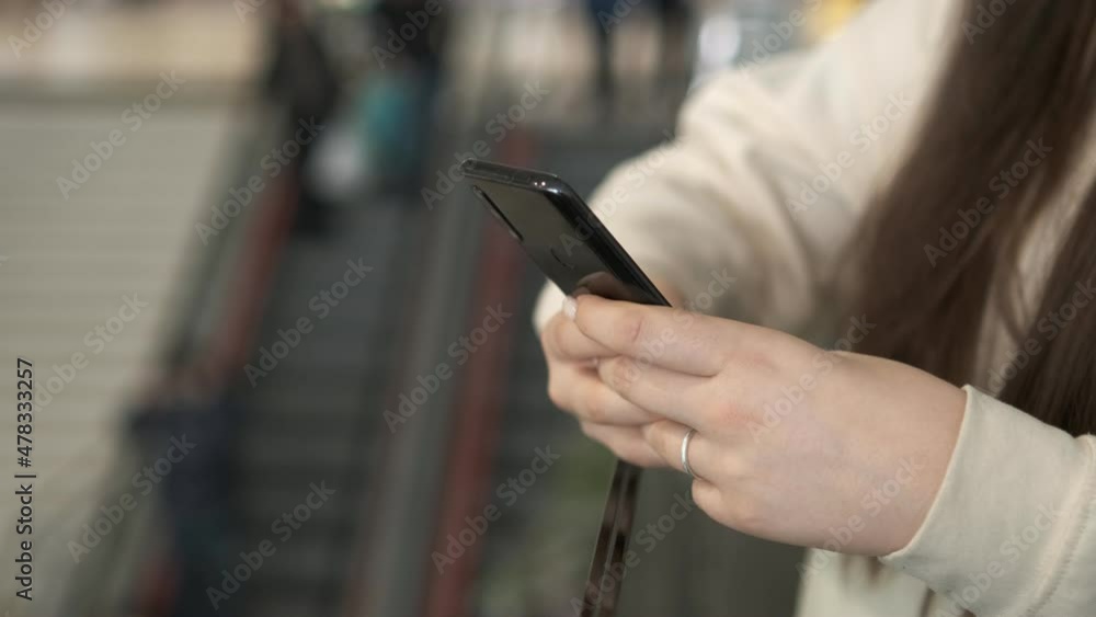 Hands of a young woman using a smartphone in a shopping mall. A young woman is holding a phone in a shopping mall in front of an escalator with people. Close up Hands of a young woman using a smartphone in a shopping mall. A young woman is holding a phone in a shopping mall in front of an escalator with people. Close up