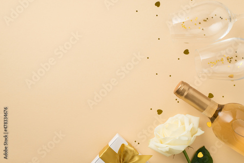 Top view photo of white rose two wineglasses with golden sequins heart shaped confetti gift box with gold bow and bottle of white wine on isolated beige background with copyspace