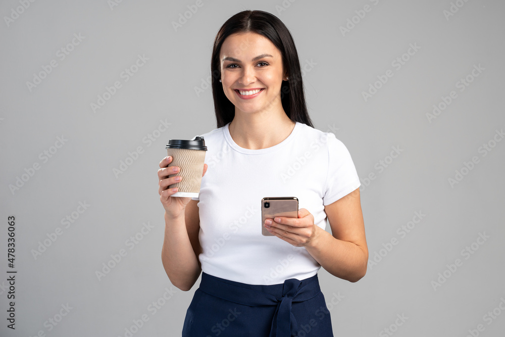 Waist up portrait view of the nice female holding cup of tasty delicious coffee in hands and smartphone isolated on grey background Waist up portrait view of the nice female holding cup of tasty delicious coffee in hands and smartphone isolated on grey background