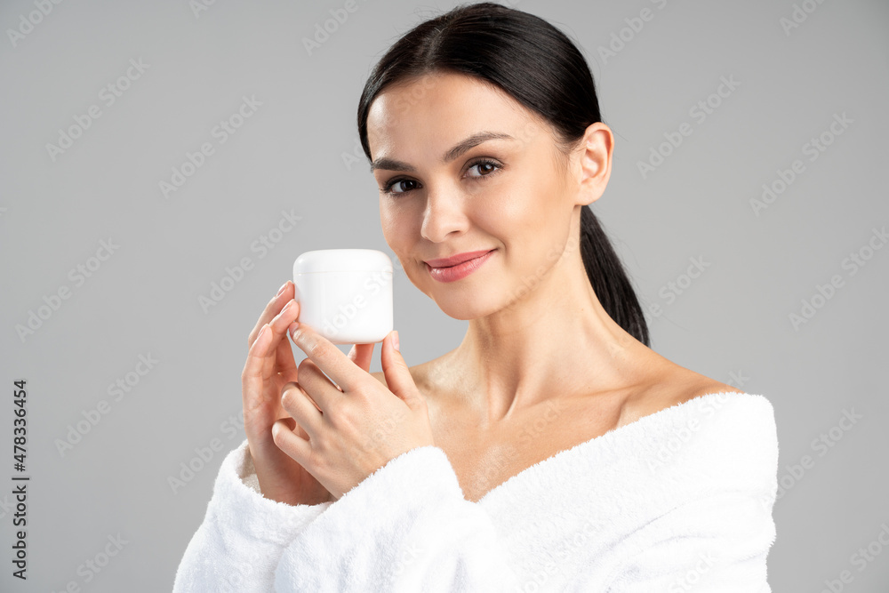 Young woman in bathrobe posing with jar of the moisturizer over isolated grey background while present a product Young woman in bathrobe posing with jar of the moisturizer over isolated grey background while present a product