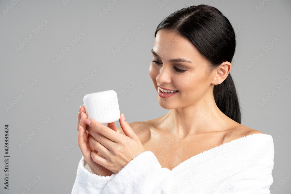 Photo portrait of caucasian brunette woman holding cream jar in hands and looking at it isolated on grey colored background Photo portrait of caucasian brunette woman holding cream jar in hands and looking at it isolated on grey colored background