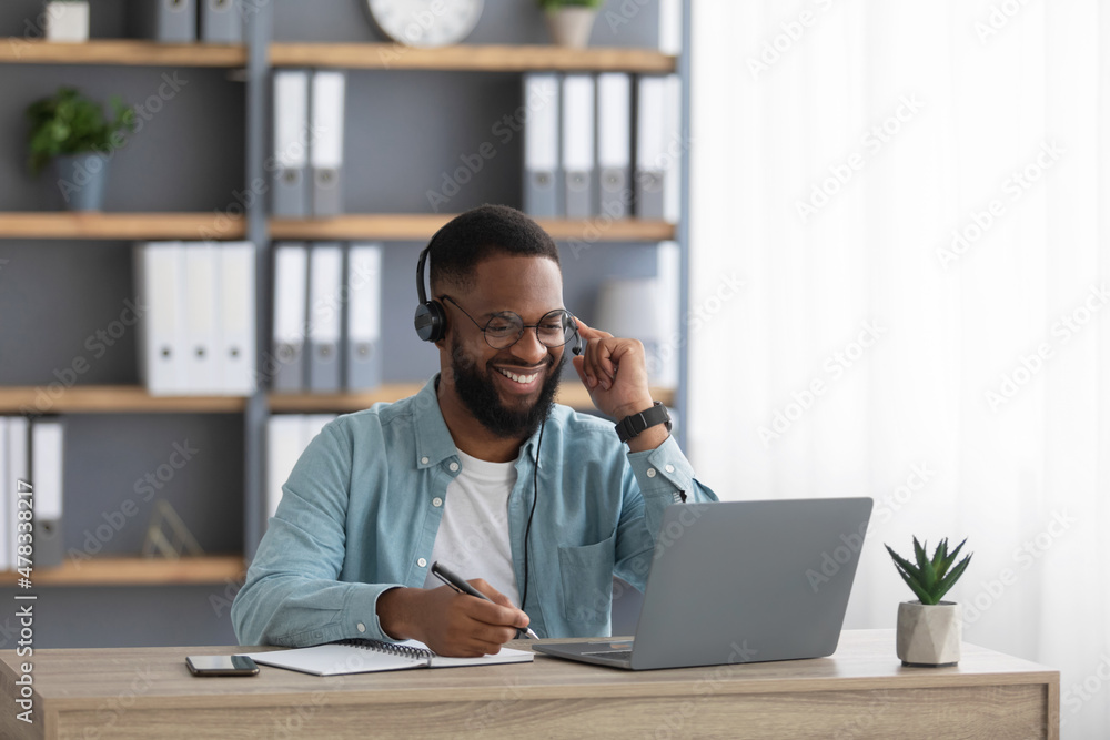 Cheerful handsome millennial african american male with beard in glasses, headphones works on computer Cheerful handsome millennial african american male with beard in glasses, headphones works on computer