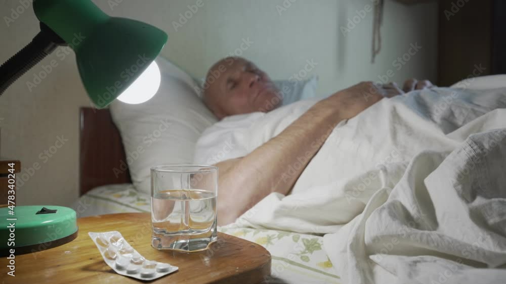 Older ill man lying in bed of hospice with pills and water on wooden nightstand. Elder human care conception. Older ill man lying in bed of hospice with pills and water on wooden nightstand. Elder human care conception.