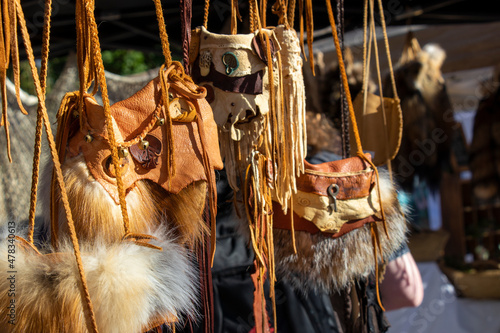Handmade purses in leather and fur with braided straps for sale, some with bells, bone buttons, and fringe, at an outdoor market.