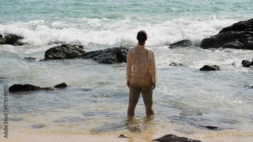 Man stand in water at sea shore, look to breaking wave rushing up between rough stones. He raise and stretch out hands, make symbolic gesture. Take in nature concept photo