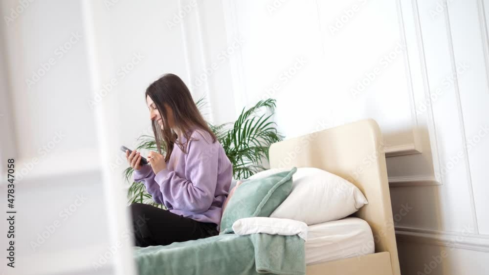 Woman sits on a bed in a bedroom, communicates using a smartphone and rejoices at the good news. Leisure at home, Staying at home, Beautiful woman, happy woman, good news. Woman sits on a bed in a bedroom, communicates using a smartphone and rejoices at the good news. Leisure at home, Staying at home, Beautiful woman, happy woman, good news.