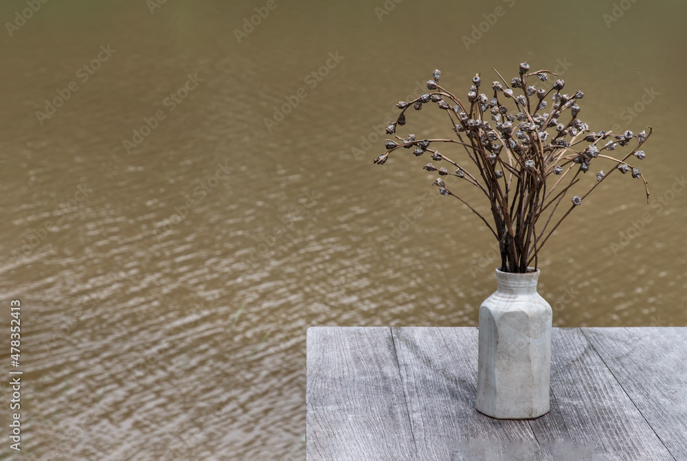 Vintage tone image of Dried flowers in white vase for decoration purpose on old wooden table with river view in background. Space for text, Selective focus.