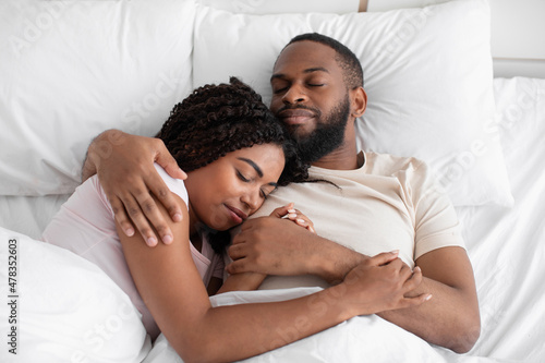 Happy young black husband hugs his wife and lie on bed on soft pillow under white blanket in bedroom
