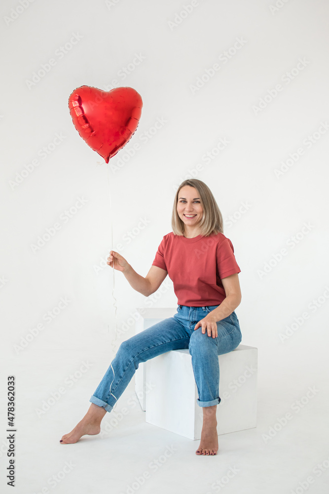 Millennial girl in blue jeans and a red T-shirt with a heart-shaped balloon on a white background. Happy Valentine's Day Millennial girl in blue jeans and a red T-shirt with a heart-shaped balloon on a white background. Happy Valentine's Day