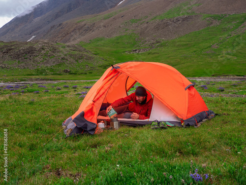 Solo trekking. Camping on a summer green rocky high-altitude plateau. Man in a orange tent is preparing food. Peace and relaxation in nature.