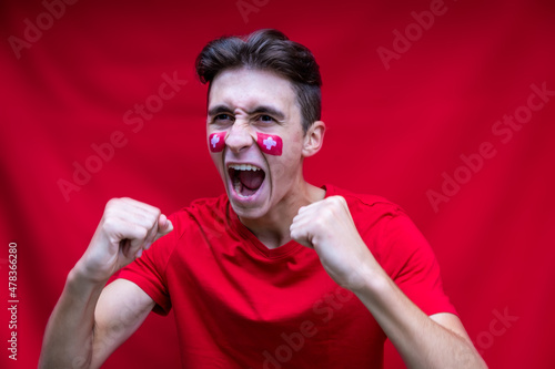 Swiss football fan shouting for his national team, isolated on red background. Flag painted over face