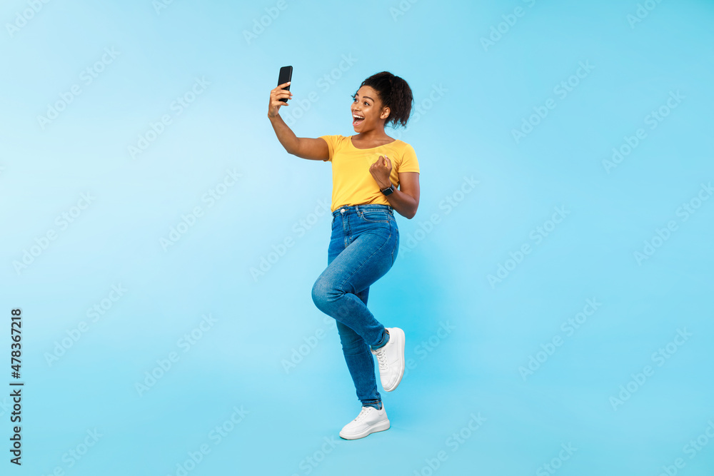 Full length of emotional young black woman looking at smartphone screen, making YES gesture, jumping in excitement Full length of emotional young black woman looking at smartphone screen, making YES gesture, jumping in excitement