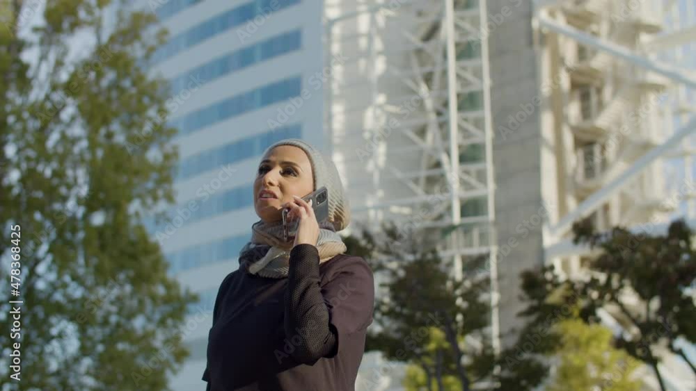 Beautiful Arab woman in hijab talking on phone outdoor. Medium shot of cheerful woman standing in background of skyscraper, having interesting conversation with boyfriend. Communication concept Beautiful Arab woman in hijab talking on phone outdoor. Medium shot of cheerful woman standing in background of skyscraper, having interesting conversation with boyfriend. Communication concept