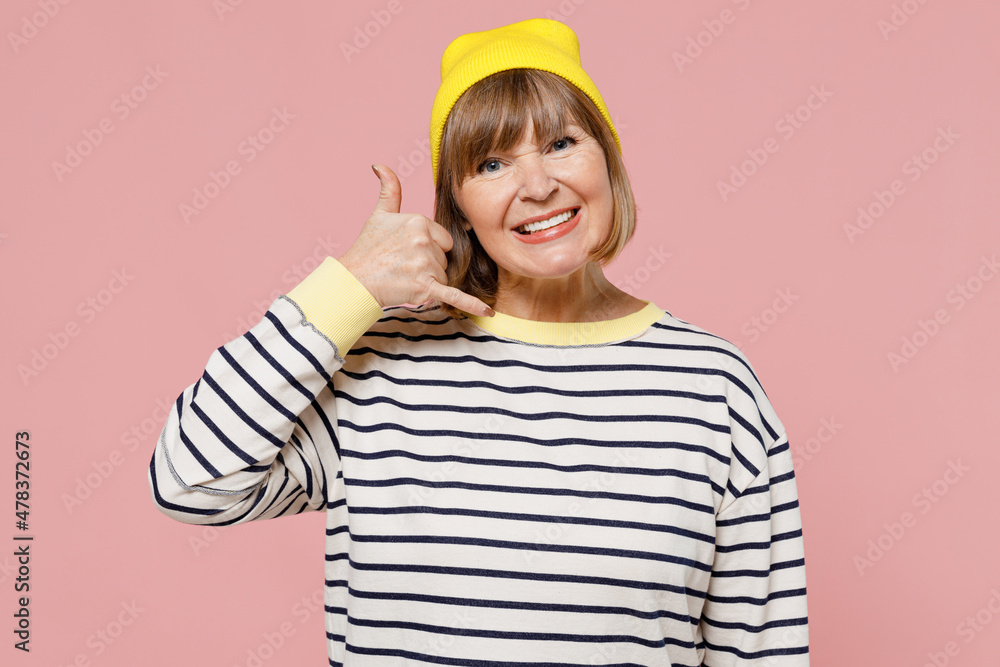 Elderly smiling stylish woman 50s wearing striped shirt yellow hat doing phone gesture like says call me back isolated on plain pastel light pink background studio portrait. People lifestyle concept. Elderly smiling stylish woman 50s wearing striped shirt yellow hat doing phone gesture like says call me back isolated on plain pastel light pink background studio portrait. People lifestyle concept.
