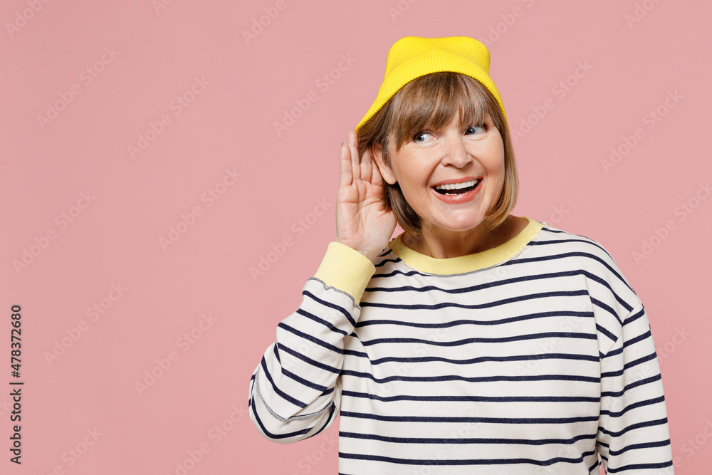 Elderly curious stylish woman 50s wearing striped shirt yellow hat try to hear you overhear listening intently isolated on plain pastel light pink background studio portrait. People lifestyle concept.