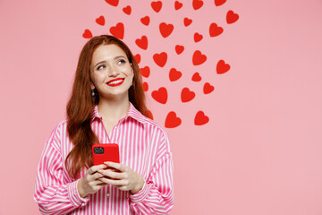 Young smiling dreamful woman 20s wearing casual striped shirt use mobile cell phone chat isolated on pink background studio, red hearts on wall Valentine's Day birthday holiday party dating concept