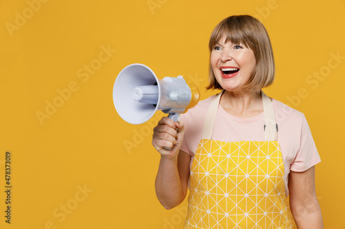 Elderly housekeeper housewife woman 50s in orange apron hold scream megaphone announces discounts sale Hurry up isolated plain on yellow background studio portrait. People household lifestyle concept.
