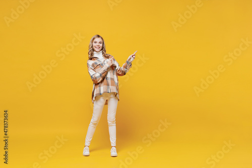 Bild auf Leinwand Full body little blonde smiling kid girl 13-14 years wearing checkered shirt point index finger aside on copy space area isolated on plain yellow background studio portrait