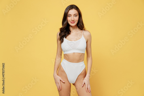 Smiling skinny sexy lovely attractive young brunette woman 20s in white underwear with beautiful perfect fit body standing posing looking camera isolated on plain yellow background studio portrait