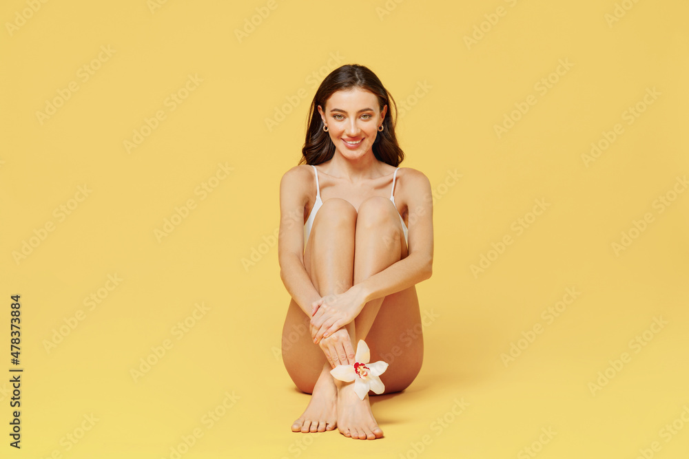 Full body smiling young nice beautiful brunette woman 20s wearing white underwear sitting on floor hold flower isolated on plain yellow color background studio portrait. People female beauty concept. Full body smiling young nice beautiful brunette woman 20s wearing white underwear sitting on floor hold flower isolated on plain yellow color background studio portrait. People female beauty concept.