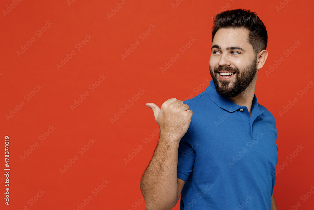 Fototapeta premium Young happy caucasian man 20s wear basic blue t-shirt looking camera point thumb finger aside on workspace area mock up isolated on plain orange background studio portrait. People lifestyle concept.