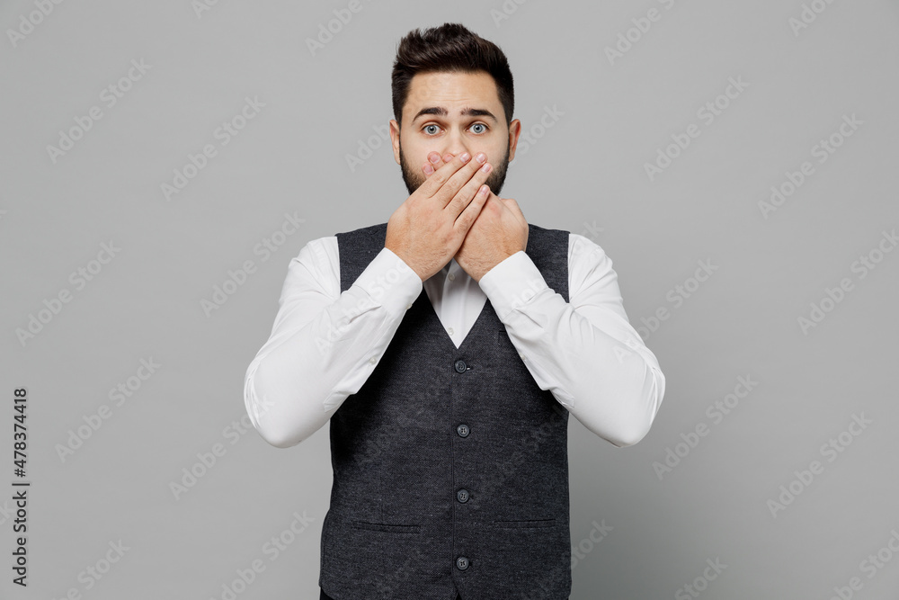 Young scared barista male waiter butler man 20s wear white shirt vest elegant uniform work at cafe cover mouth with hand isolated on plain grey background studio portrait. Restaurant employee concept. Young scared barista male waiter butler man 20s wear white shirt vest elegant uniform work at cafe cover mouth with hand isolated on plain grey background studio portrait. Restaurant employee concept.