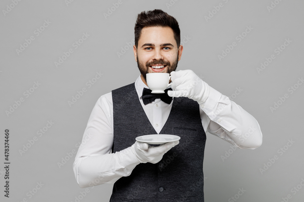 Young barista male waiter butler man 20s in white shirt vest elegant uniform work at cafe drink coffee hold cup with hot beverage isolated on plain grey background studio Restaurant employee concept Young barista male waiter butler man 20s in white shirt vest elegant uniform work at cafe drink coffee hold cup with hot beverage isolated on plain grey background studio Restaurant employee concept
