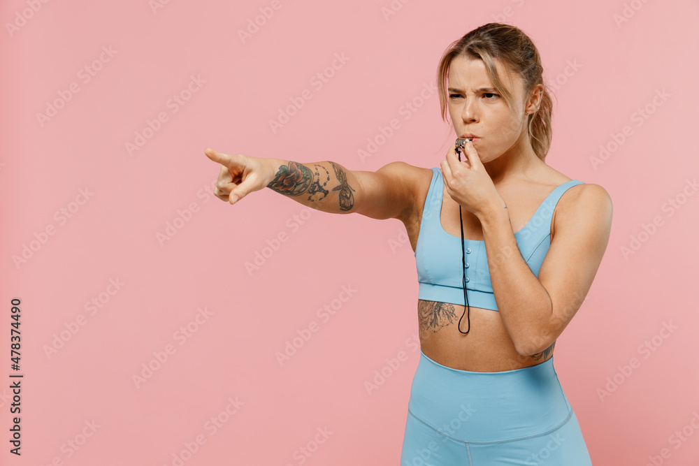Young strong sporty athletic fitness trainer instructor woman wear blue tracksuit spend time in home gym whistle point finger aside isolated on plain pink background. Workout sport motivation concept. Young strong sporty athletic fitness trainer instructor woman wear blue tracksuit spend time in home gym whistle point finger aside isolated on plain pink background. Workout sport motivation concept.