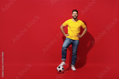 Fototapeta Naklejka Na Ścianę i Meble -  Full size body length happy young bearded man football fan in yellow t-shirt cheer up support favorite team look camera hold foot on soccer ball isolated on plain dark red background studio portrait.