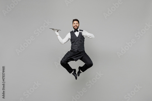 Full body fun young male waiter butler man 20s wear white shirt vest elegant uniform work at cafe jump high point finger on metal tray isolated on plain grey background . Restaurant employee concept.