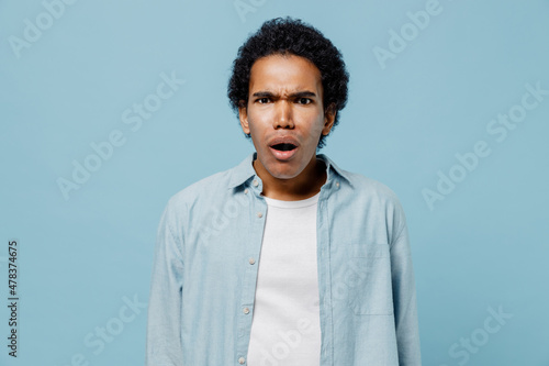 Shocked surprised stupefied frightened upset young black curly man 20s years old wear white shirt looking camera keeping mouth wide open isolated on plain pastel light blue background studio portrait