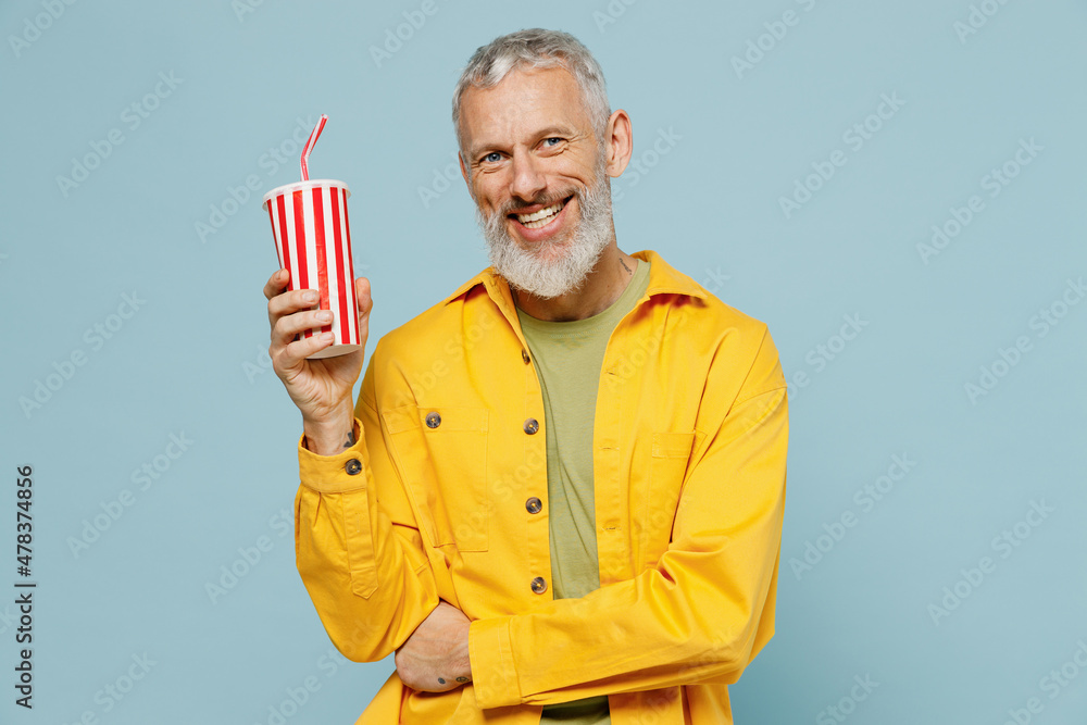 Elderly happy gray-haired mustache bearded man 50s wear yellow shirt hold cup of soda cola fizzy water beverage isolated on plain pastel light blue background studio portrait People lifestyle concept