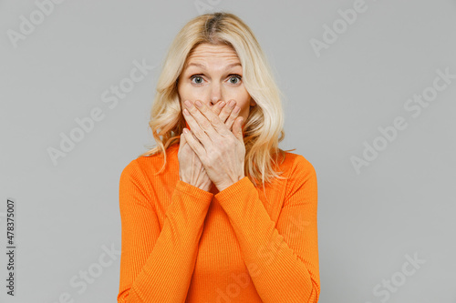 Elderly shocked surprised scared astonished blonde caucasian woman 50s wear orange turtleneck cover mouth with hand isolated on plain grey color background studio portrait. People lifestyle concept. photo