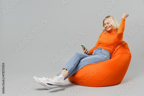 Full body elderly smiling blonde woman 50s in orange turtleneck sit in bag chair hold in hand use mobile cell phone do winner gesture isolated on plain grey background studio People lifestyle concept.