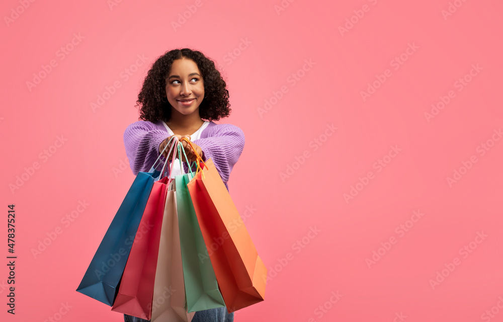 Cool shopping offer. Beautiful young black woman showing shopper bags at camera, smiling on pink studio background