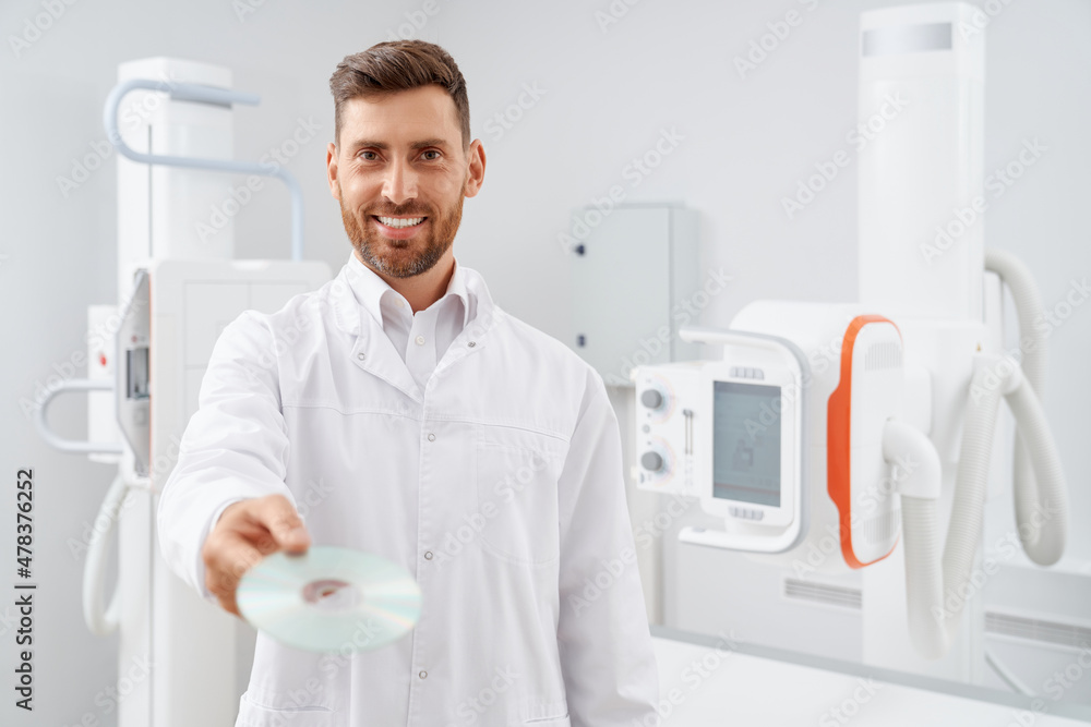 Handsome radiologist with beard given showing at camera silver CD plate with result after diagnostic. Professional doctor working with ultrasound equipment holding electronic information about health. Handsome radiologist with beard given showing at camera silver CD plate with result after diagnostic. Professional doctor working with ultrasound equipment holding electronic information about health.