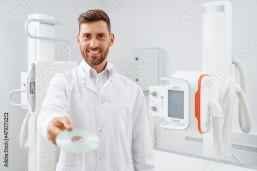Handsome radiologist with beard given showing at camera silver CD plate with result after diagnostic. Professional doctor working with ultrasound equipment holding electronic information about health.