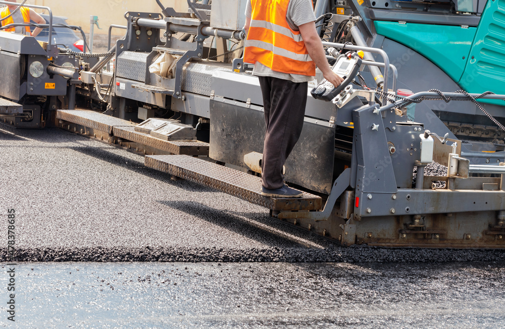 A road worker operates a tracked asphalt paver while paving fresh asphalt on the road on a summer day. A road worker operates a tracked asphalt paver while paving fresh asphalt on the road on a summer day.