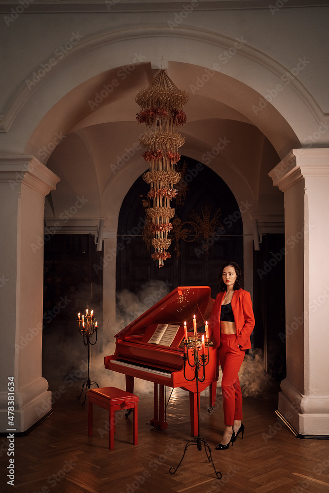 Fototapeta premium The girl plays the red piano. She is wearing a stylish and trendy red suit. Evening, candles.