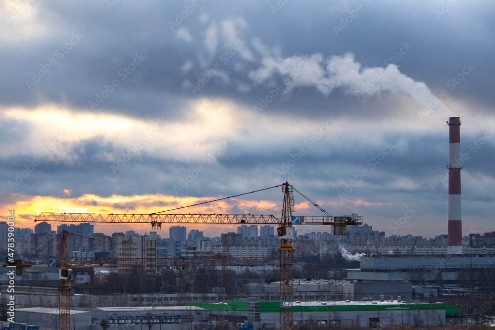 A cold autumn sunrise with a line of lumen from the sun over the houses on the outskirts of the metropolis landscape with a smoking pipe in Russia. A smoking pipe and two cranes in the foreground. A cold autumn sunrise with a line of lumen from the sun over the houses on the outskirts of the metropolis landscape with a smoking pipe in Russia. A smoking pipe and two cranes in the foreground.
