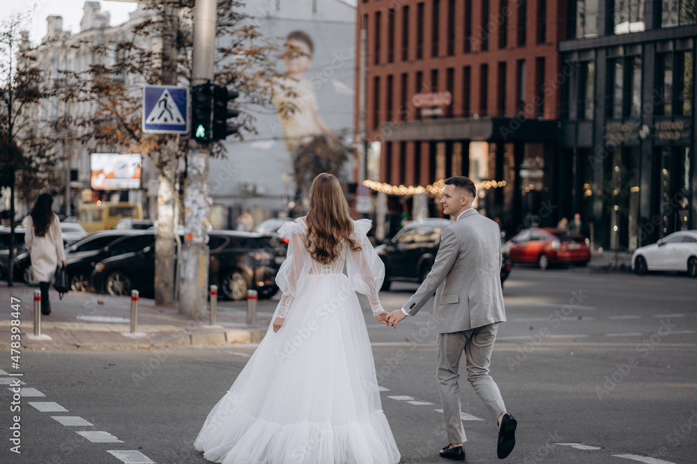 Beautiful stylish European bride and groom walking on the summer streets. Woman holds man's hand walking with him on the city
