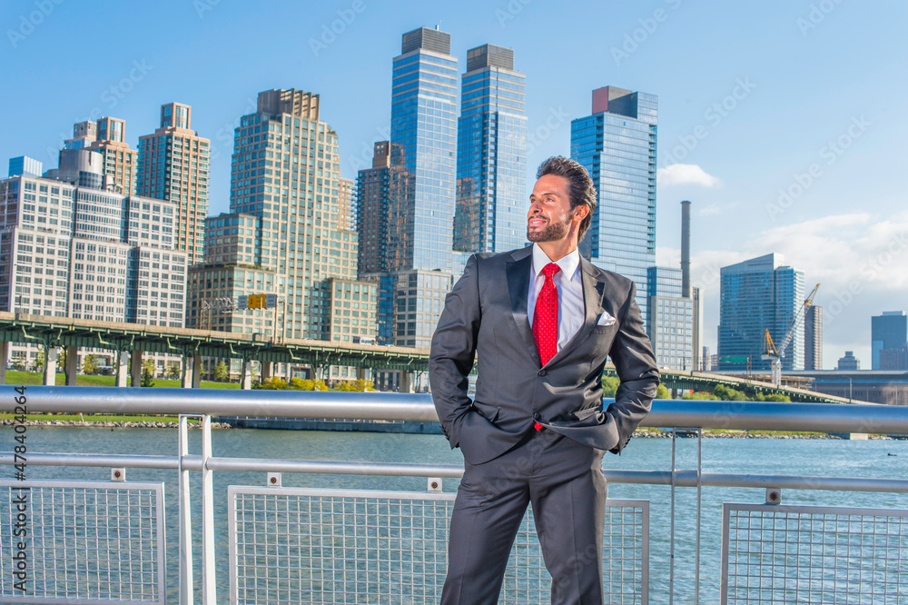 Dressing is a black suit, a red tie, hands in pockets, a sexy, middle age businessman with mustache and beard is standing in the front of a business district, confidently looking forward. Dressing is a black suit, a red tie, hands in pockets, a sexy, middle age businessman with mustache and beard is standing in the front of a business district, confidently looking forward.