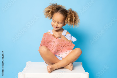 Cute little girl sitting on a table writing in notebook isolated over blue background. Back to school concept.