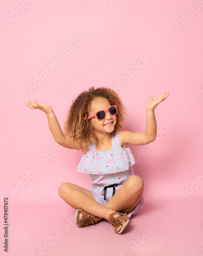 Cute little girl wearing sunglasses sitting on the floor with hands up over pink background. © Danko