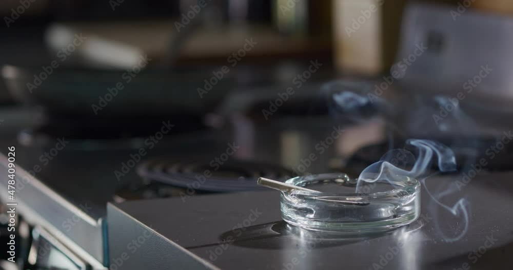 Interior detail shot of ashtray that has a lit marijuana CBD pre-roll joint, cigarette with smoke rising sitting in kitchen next to stove top Interior detail shot of ashtray that has a lit marijuana CBD pre-roll joint, cigarette with smoke rising sitting in kitchen next to stove top